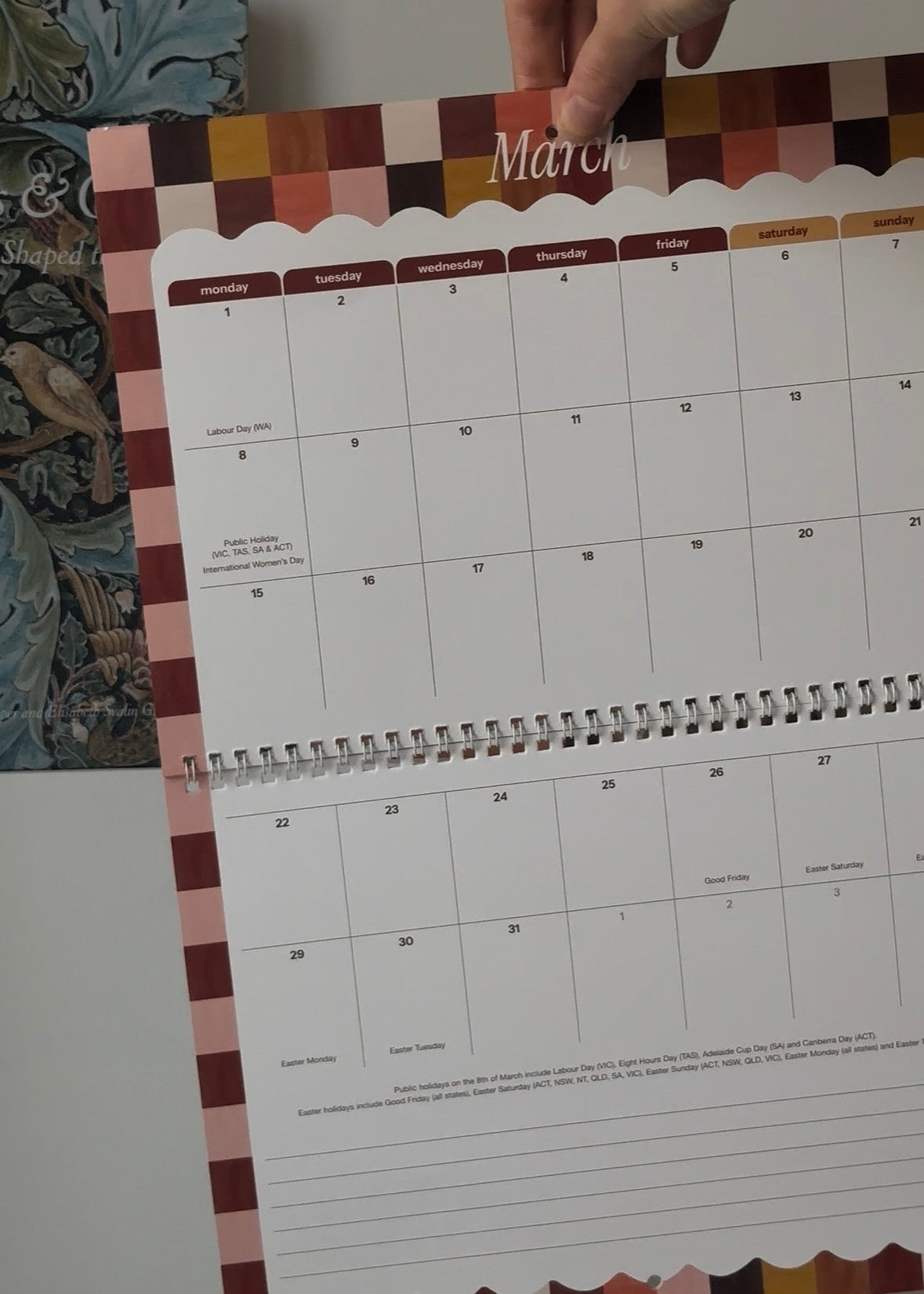 A hand holding open a spiral-bound monthly calendar page showing the month of March, with a patterned border in shades of pink, brown, and beige.