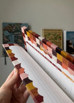 A hand holding an open monthly calendar or planner with lined pages and a multicolor patterned cover in a home setting.