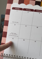 A close-up of a spiral-bound monthly calendar page showing days of the week, held by a hand. The calendar has white pages with dark red headers for each day and black printed text for holidays.