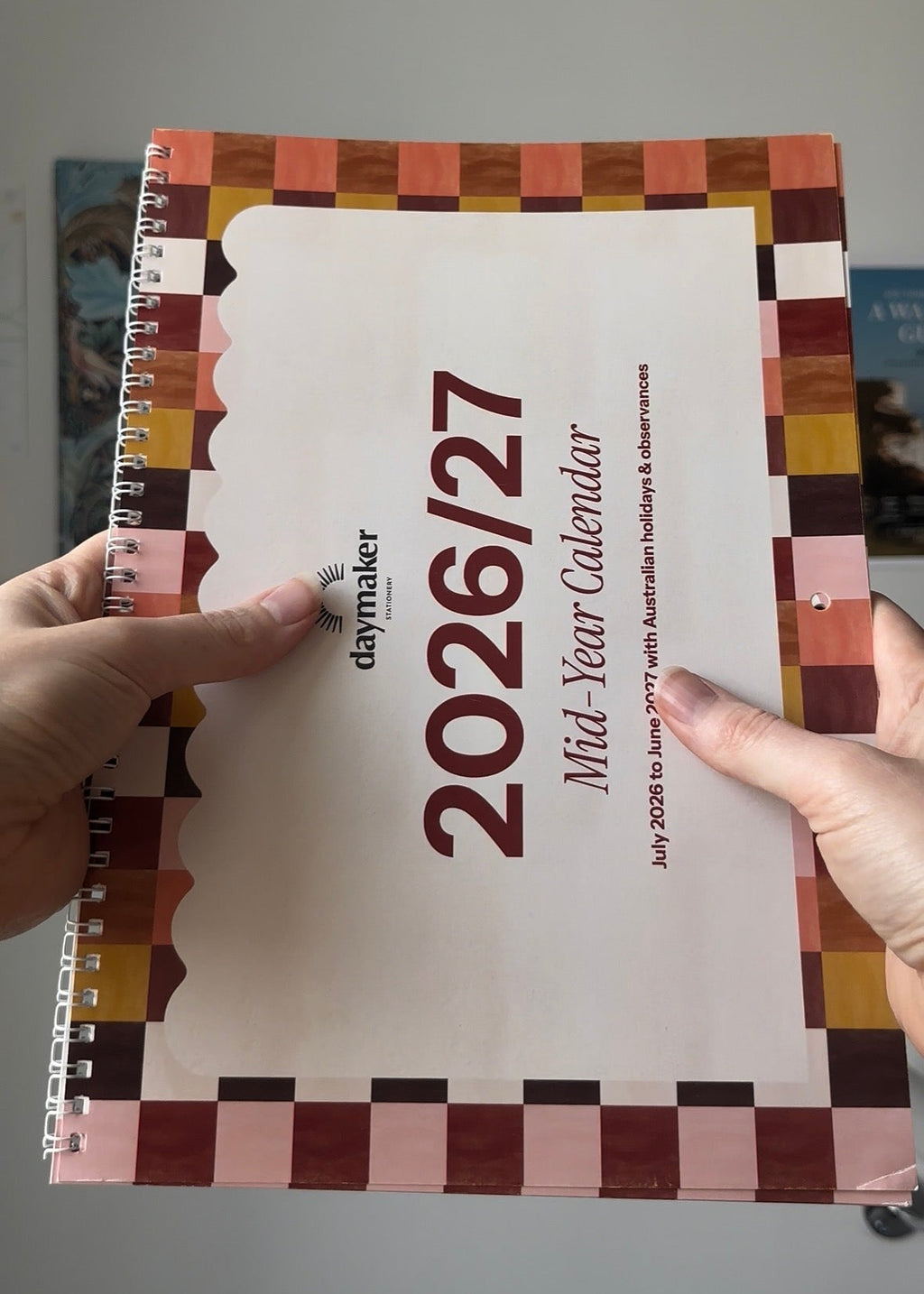 A person holds a Daymaker 2026 Mid-Year Calendar with a cream cover and a geometric multicolor border in shades of pink, brown, orange, and yellow.
