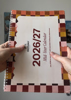 A person holds a Daymaker 2026 Mid-Year Calendar with a cream cover and a geometric multicolor border in shades of pink, brown, orange, and yellow.