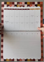 A weekly planner page from a spiral-bound desk calendar, showing the February layout with a multicolor checkered border and a blank lined section below, held open by hands.