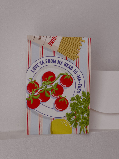 A square greeting card featuring a retro-style illustration of fresh tomatoes on a vine, spaghetti, parsley, and a lemon slice, with the text 'LOVE YA FROM MA HEAD TO-MA TOES' on a striped background. The card is displayed standing upright against a neutral light gray wall.
