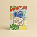 A greeting card featuring a colourful illustration of a tin of sardines / fish with the text 'Glad We're Tight' surrounded by mediterranean ingredients like tomatoes, pasta, parsley, and lemons, set against a light blue background. The card is depicted standing upright on a warm beige surface with a matching beige background. This card is suitable for Father's Day, Mother's Day, Valentines Day and many other affectionate and grateful occasions.