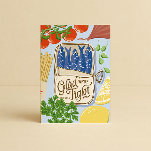 A greeting card featuring a colourful illustration of a tin of sardines / fish with the text 'Glad We're Tight' surrounded by mediterranean ingredients like tomatoes, pasta, parsley, and lemons, set against a light blue background. The card is depicted standing upright on a warm beige surface with a matching beige background. This card is suitable for Father's Day, Mother's Day, Valentines Day and many other affectionate and grateful occasions.