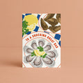 A vertical greeting card featuring an illustration of oysters and lemons, with the text 'TO A SHUCKING GREAT DAD' in red lettering curved around the edge of the plate, photographed standing against a solid peach background. 